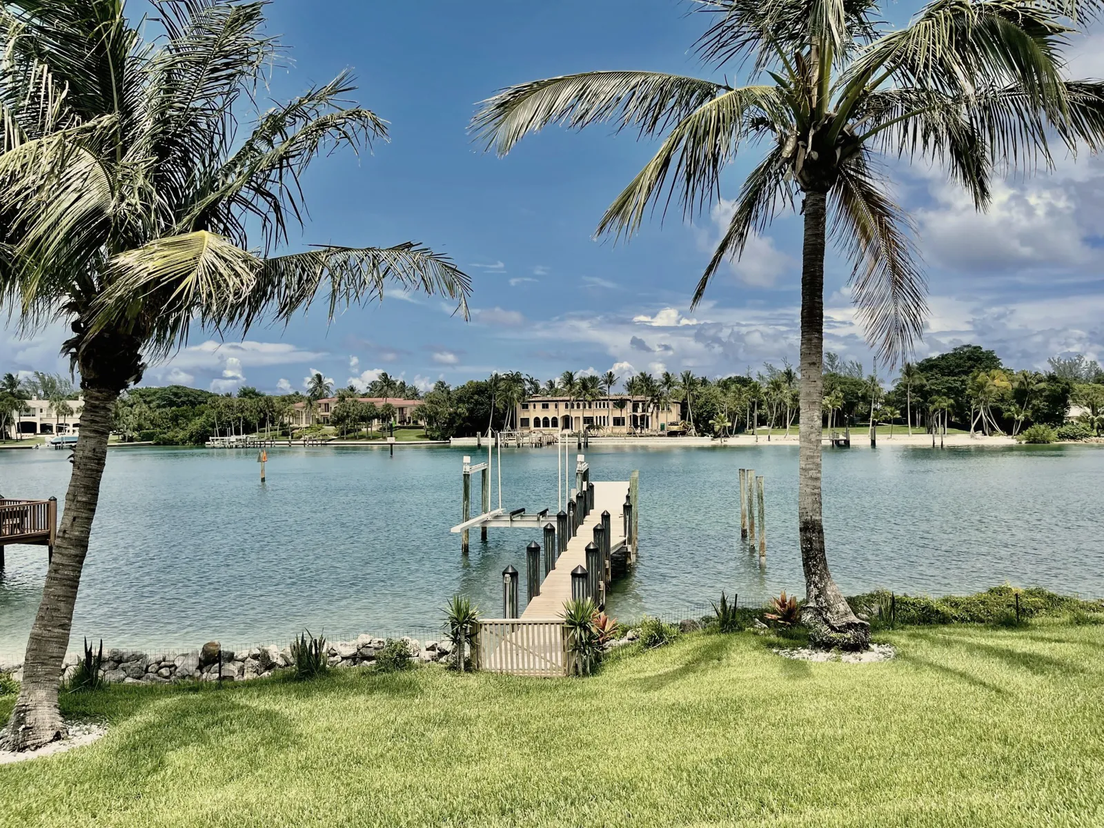 Private dock reaching into calm blue water between two palm trees, with large homes across the water