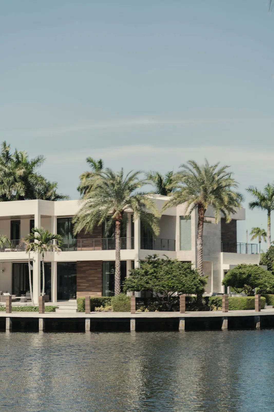 White modern waterfront home behind a seawall, with palm trees and a pale blue sky