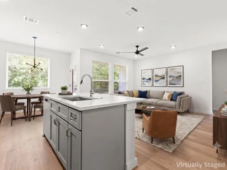 Virtually staged open-concept kitchen, dining, and living area featuring a spacious quartz island with an undermount sink, gray shaker-style cabinetry, stainless steel appliances, recessed lighting, and light wood flooring. The kitchen flows seamlessly into a bright dining area with large windows overlooking mature trees and a comfortable living room with contemporary furnishings, creating an ideal space for entertaining and everyday living.